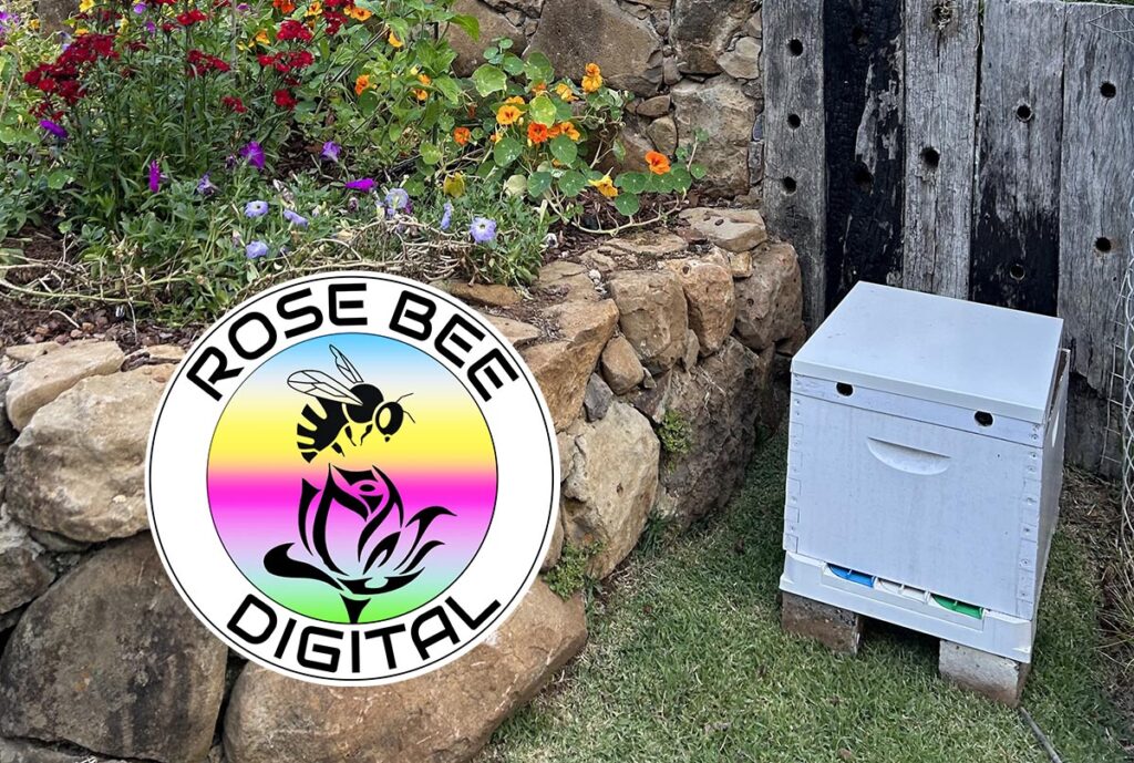 The Rose Bee beehive is located next to a colourful, raised flowerbed garden, beside a rustic rock wall. The Rose Bee Digital logo has a rainbow coloured background.