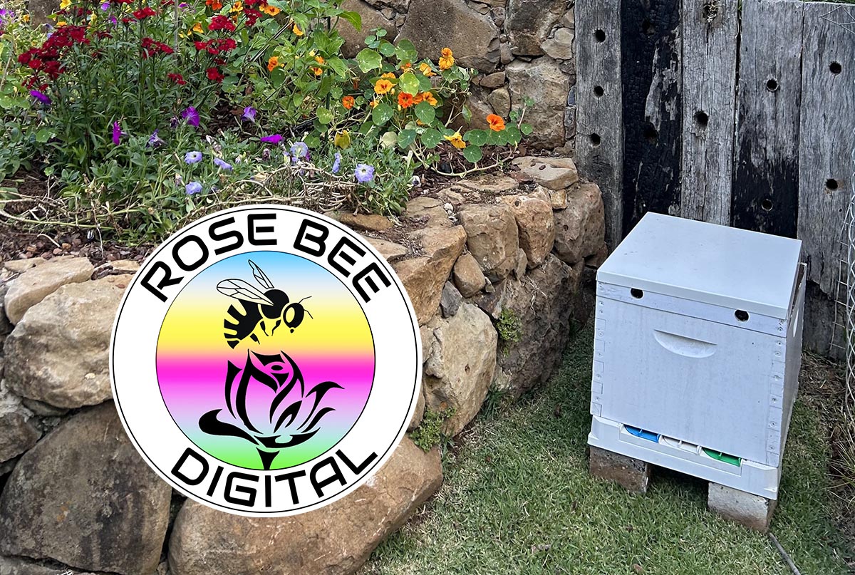 The Rose Bee beehive is located next to a colourful, raised flowerbed garden, beside a rustic rock wall. The Rose Bee Digital logo has a rainbow coloured background.